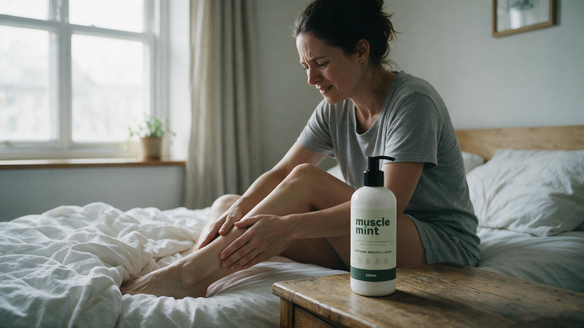 A woman sitting on the edge of her bed reaching down to a sore calf the morning after training, with a Muscle Mint 250ml bottle on the bedside table