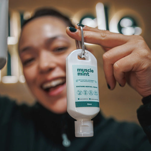 A person holding a small white hand sanitizer bottle labeled 'muscle mint' with a metal key ring, with their face blurred in the background.