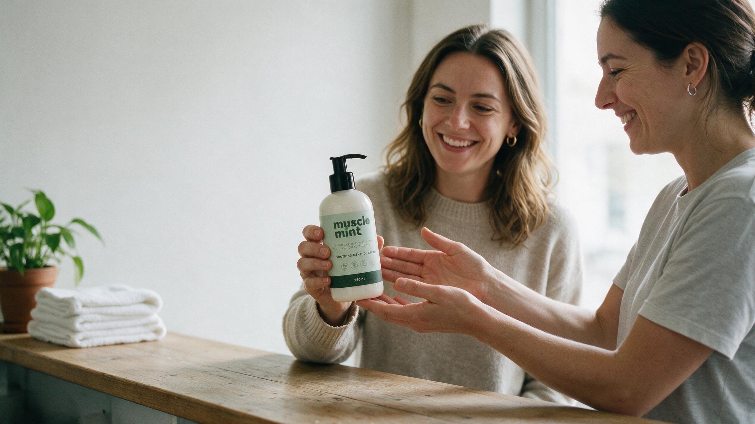 One woman handing a Muscle Mint 250ml bottle to another across a studio reception counter in daylight