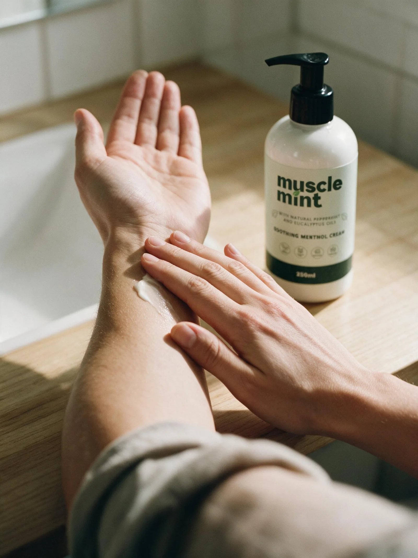 A person massaging Muscle Mint into their bare forearm at a bathroom counter, the 250ml bottle resting nearby