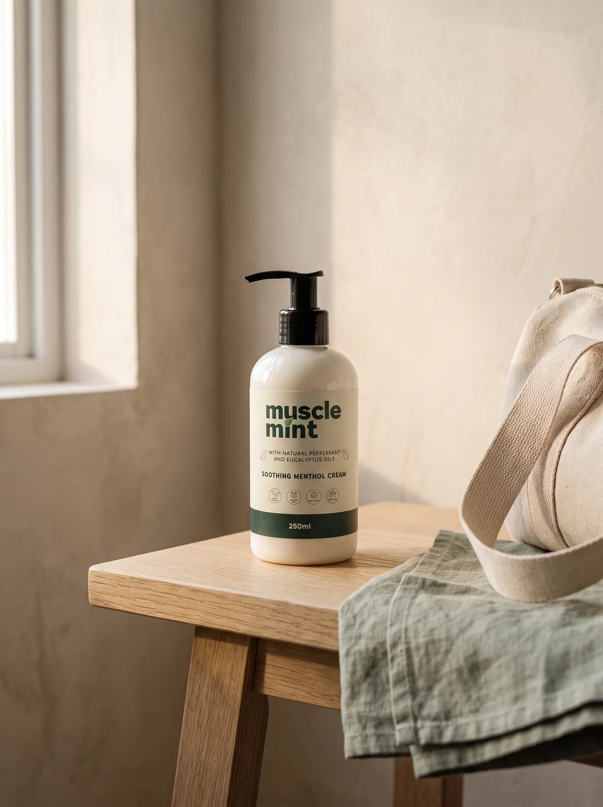 Muscle Mint 250 ml bottle on a pale oak bench beside a folded sage linen towel and a gym bag, in soft window light