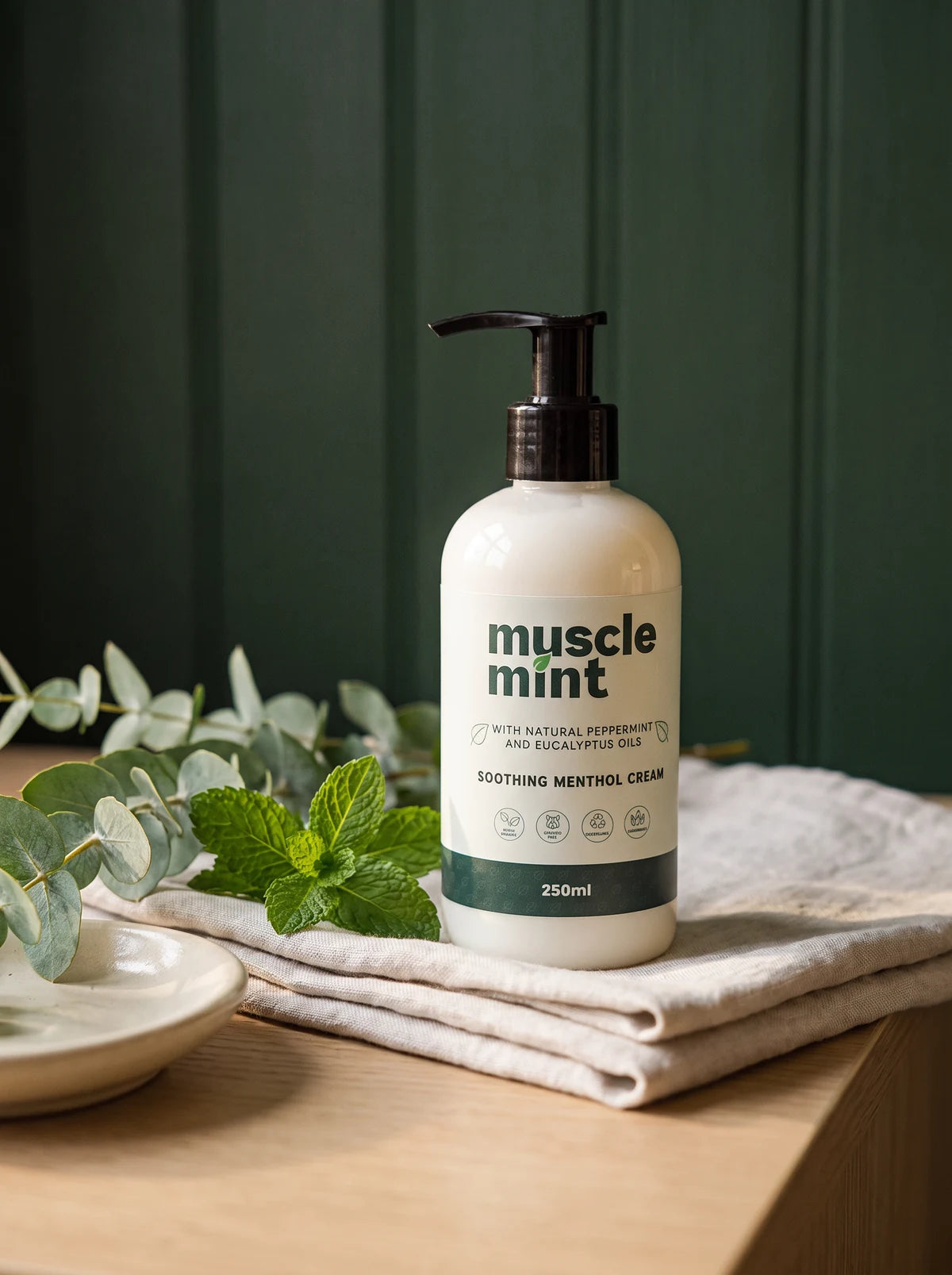 Muscle Mint 250 ml bottle on folded linen with eucalyptus and mint leaves, against deep green panelling