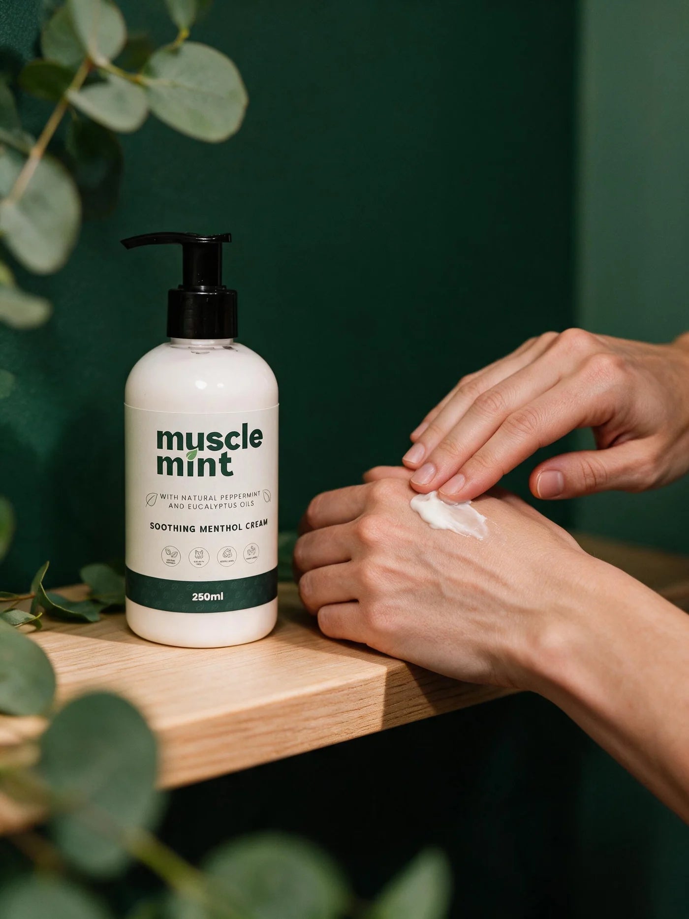 Muscle Mint 250ml bottle on a wooden shelf while cream is smoothed into the back of a hand