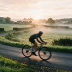 A cyclist rides a road bike along a winding country road at sunrise, with misty fields and distant houses in the background.