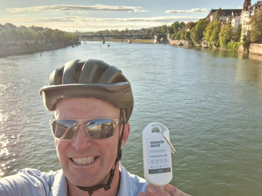 Muscle Mint 50ml travel size menthol cream held up by a cyclist in helmet and sunglasses, river and arched bridge behind, Hégenheim, France