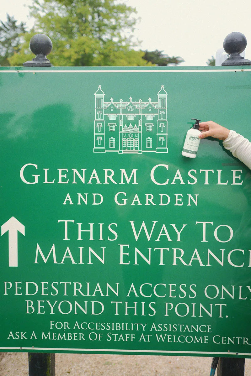 Hand holding a bottle of Muscle Mint beside the green Glenarm Castle and Garden entrance sign in Northern Ireland.