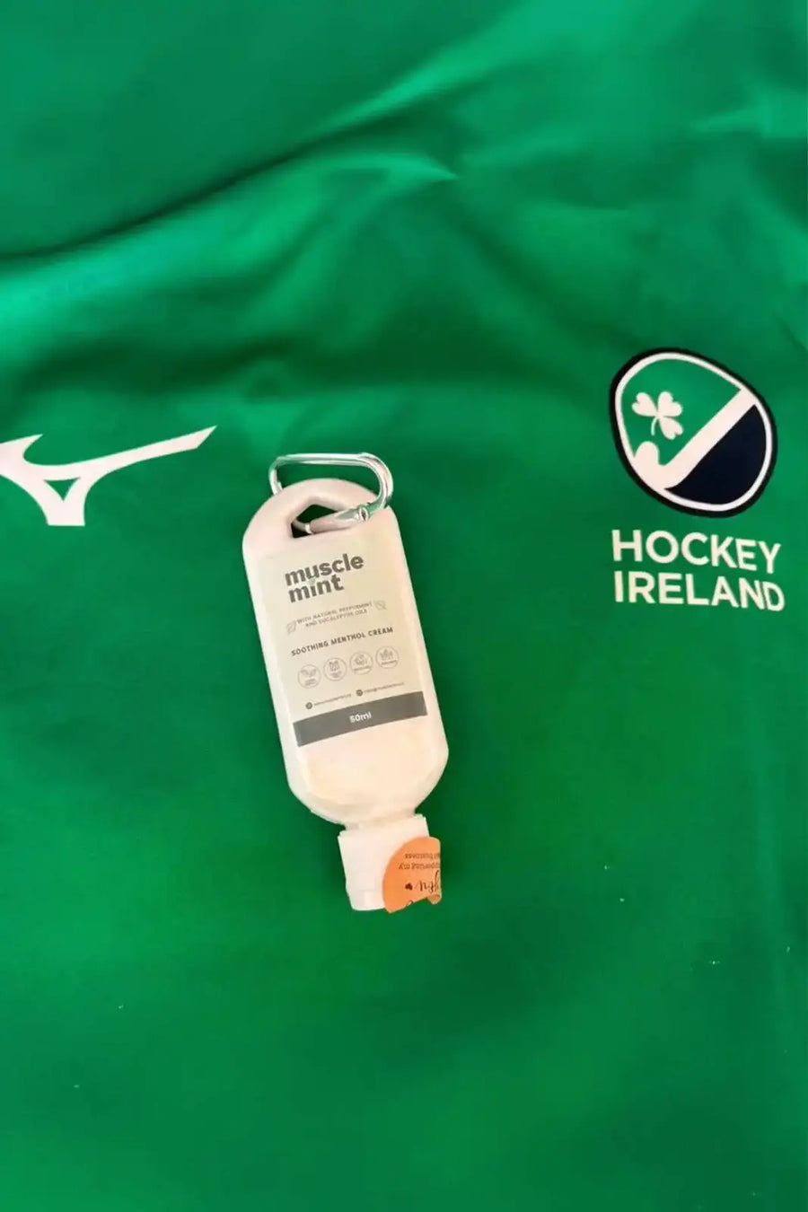 Muscle Mint 50ml travel bottle with carabiner clip resting on an Ireland Hockey jersey, packed ahead of the team's trip to Belfius Hockey Arena in Wavre, Belgium.