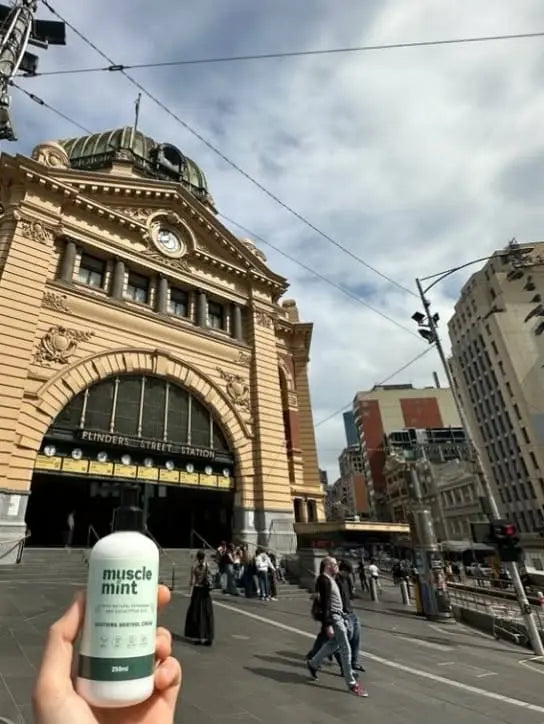 Muscle Mint 250ml menthol cream held up in front of the arched entrance and clocks of Flinders Street Station, Melbourne, Australia