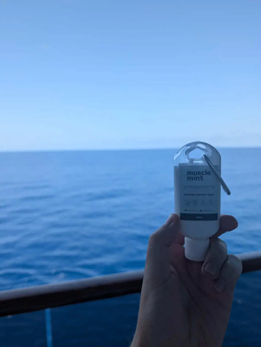 Hand holding Muscle Mint 50ml travel size menthol cream over a ship's rail with open blue water and an empty horizon behind, Balearic Sea