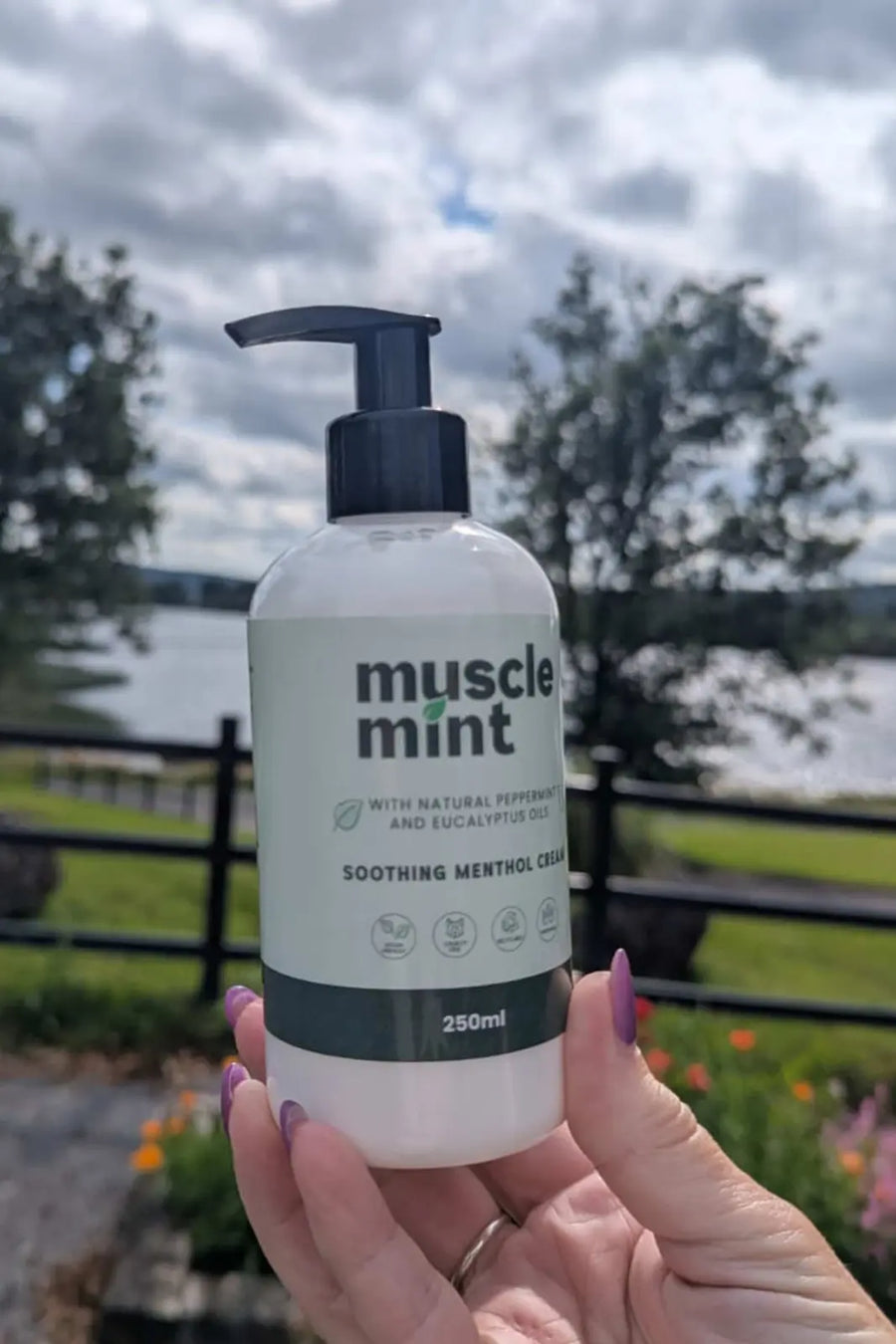 Hand holding a 250ml bottle of Muscle Mint soothing menthol cream at Lough Arrow Touring Park, Ireland, with the lake and campsite in the background