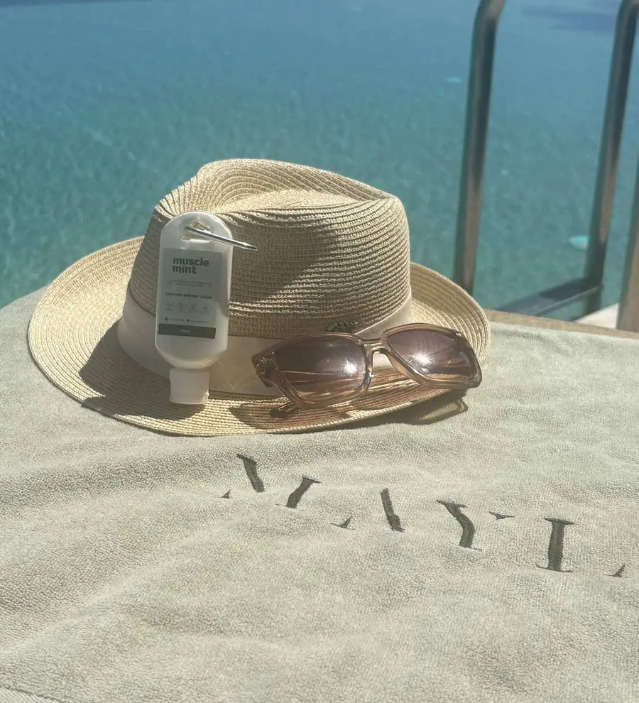 Muscle Mint travel size menthol cream clipped to a straw sun hat with sunglasses on a poolside towel at Mayia Exclusive Resort and Spa in Kiotari, Rhodes, with turquoise sea behind