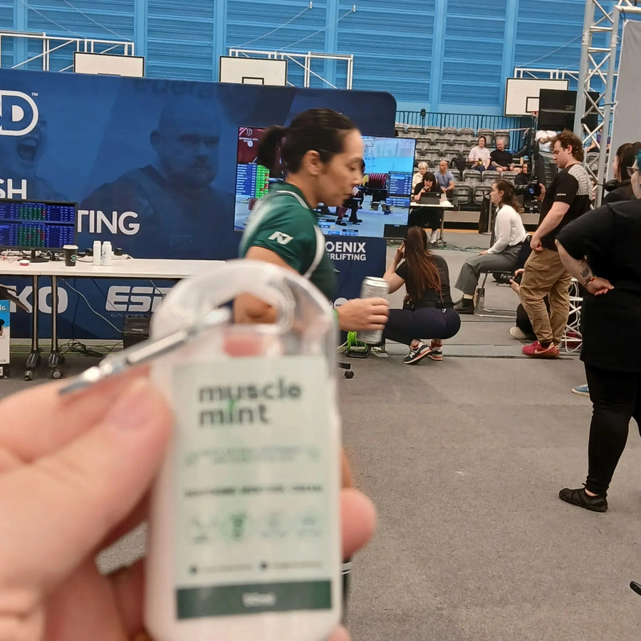 Hand holding a travel-size Muscle Mint bottle at a powerlifting championship in Edinburgh, with an athlete and competition area in the background.