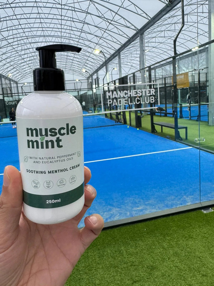 Hand holding a 250ml bottle of Muscle Mint soothing menthol cream beside a blue padel court at Manchester Padel Club.
