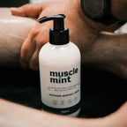 A white pump bottle of Muscle Mint Soothing Menthol Cream with a black pump, shown in a close-up lifestyle shot with a person's arm and hand in the background.
