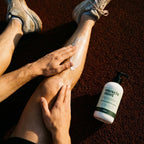 A person rubs Muscle Mint Sports Recovery Cream onto their leg after a tennis session, with the white bottle placed on the reddish-brown turf next to them.