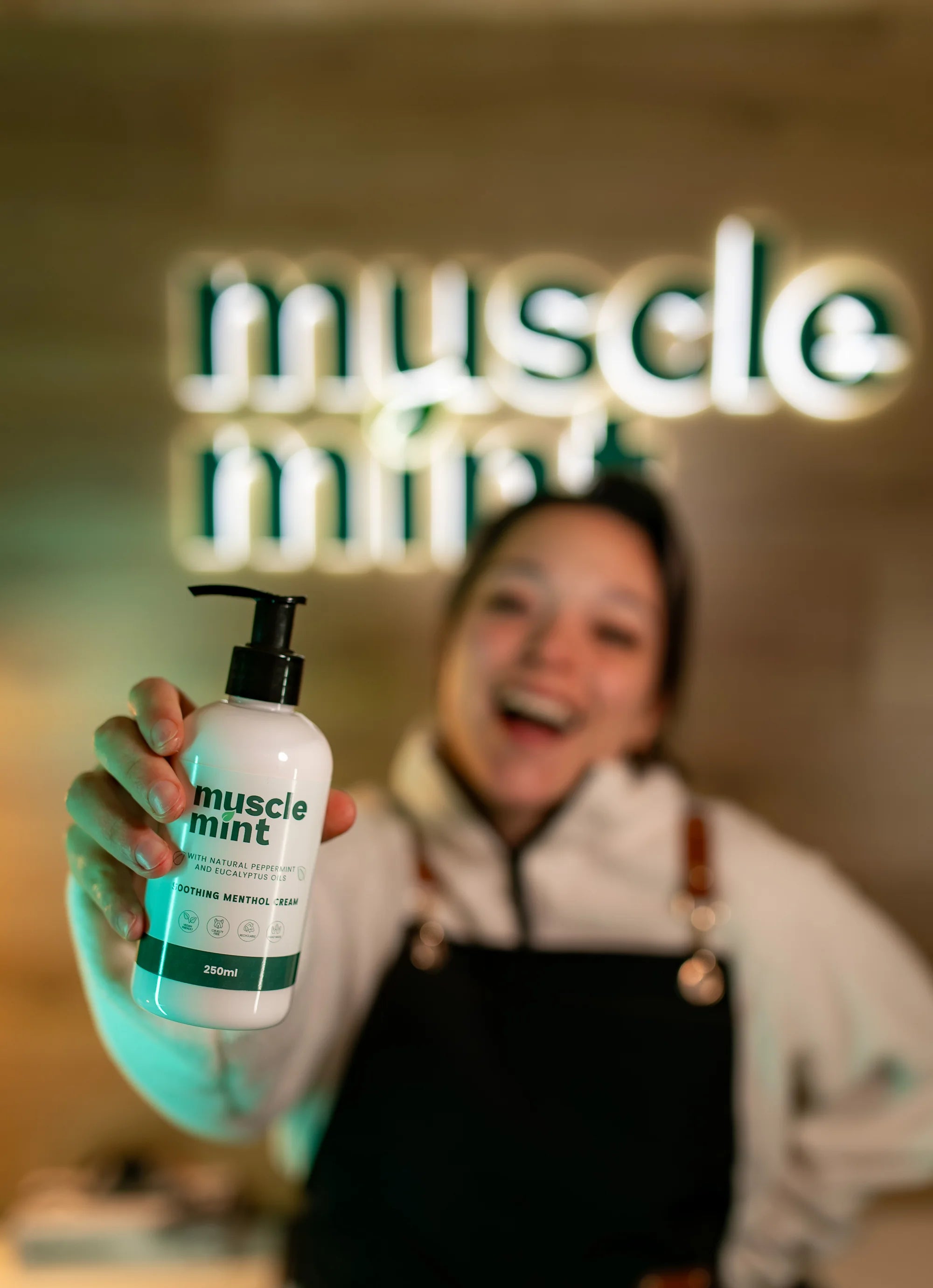 Zolene holding the Muscle Mint 250 ml bottle beneath the illuminated Muscle Mint sign