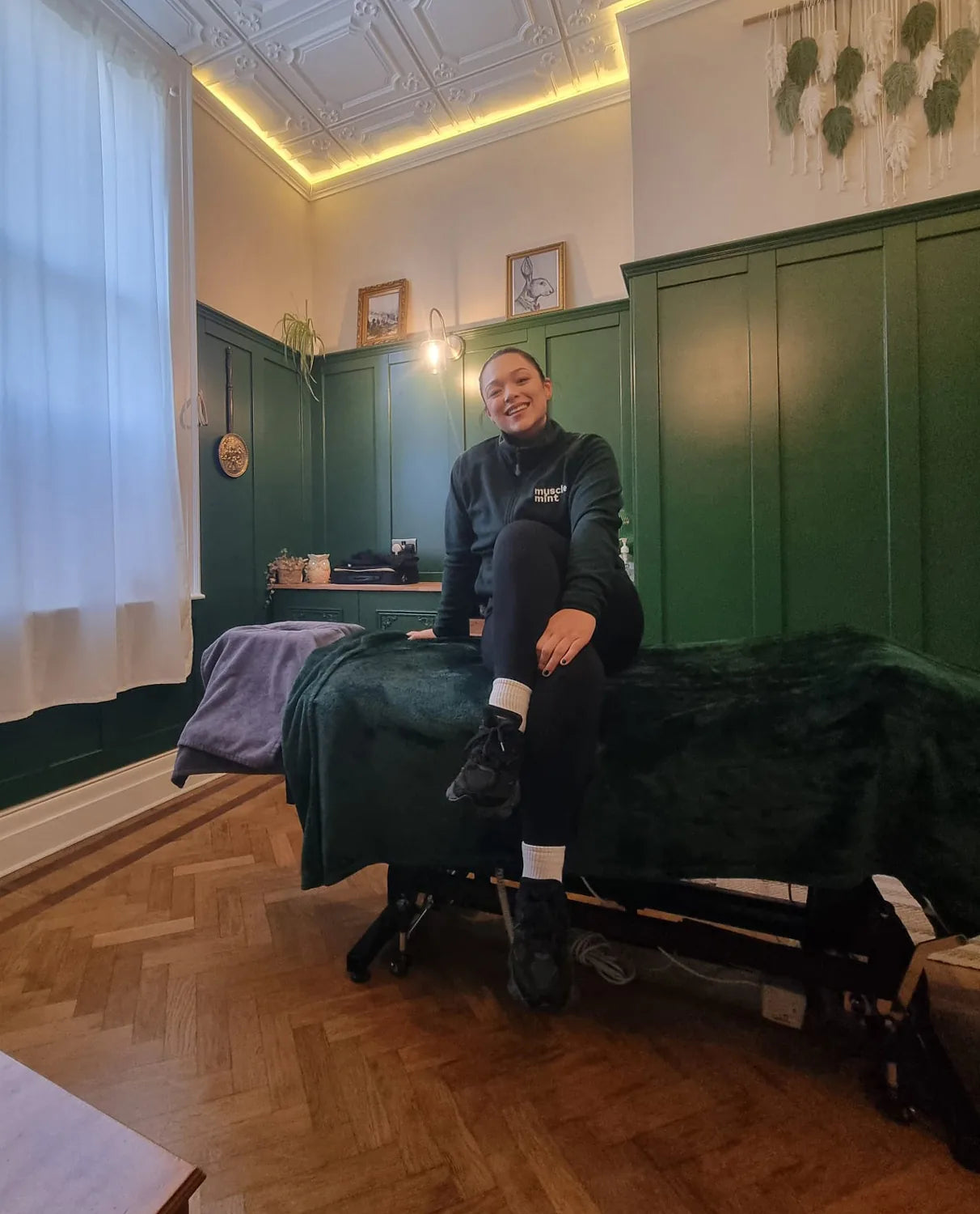 Zolene in a Muscle Mint fleece, sitting on a treatment couch in a green panelled treatment room